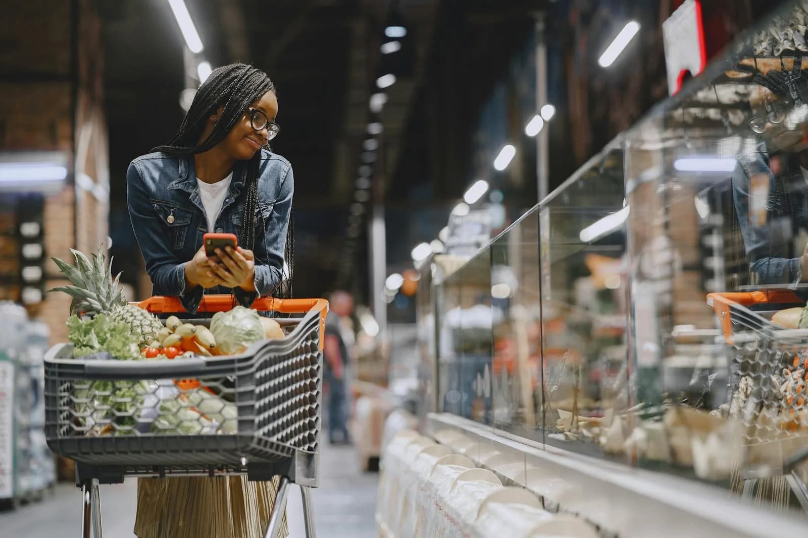 Woman in a supermarket using her smartphone while shopping with a cart full of fresh produce — connected packaging in a real shopper context